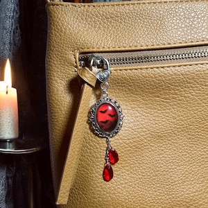 Op de afbeelding: Een bruine leren tas met een zilveren hanger bevestigd aan de rits. De hanger heeft een rode glazen cameo met een silhouet van vleermuizen tegen een donkerrode achtergrond. Drie rode, traanvormige glazen kralen hangen aan de hanger.