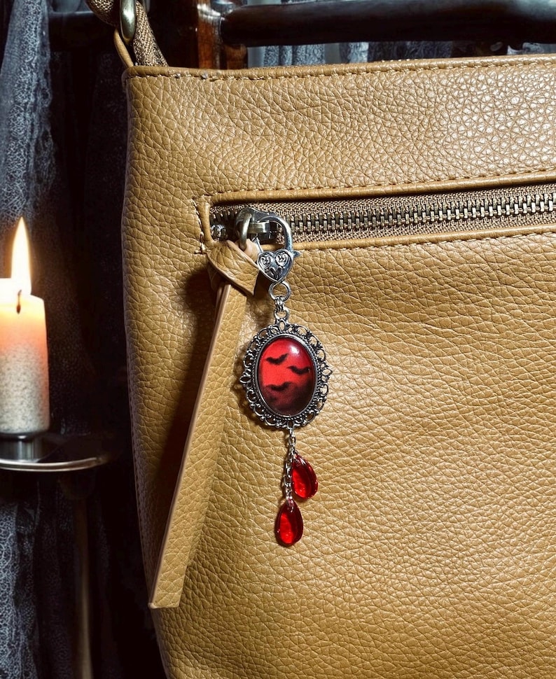 Op de afbeelding: Een bruine leren tas met een zilveren hanger bevestigd aan de rits. De hanger heeft een rode glazen cameo met een silhouet van vleermuizen tegen een donkerrode achtergrond. Drie rode, traanvormige glazen kralen hangen aan de hanger.
