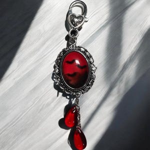 Op de afbeelding: Een zilveren bedel met een rode ovale cameo met een silhouet van vleermuizen tegen een rode en zwarte achtergrond. Twee rode traanvormige bedels hangen aan de onderkant van de cameo.