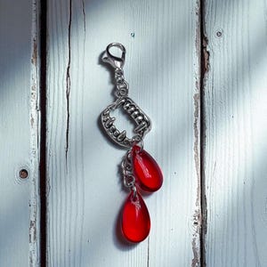 May include: A silver-colored charm featuring a set of vampire fangs and two red teardrop-shaped pendants. The charm is attached to a clasp, and the pendants are suspended by small chains. The background is a white wooden surface.
