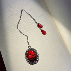 May include: A silver bookmark with a red and black oval charm featuring a silhouette of bats against a sunset. The charm is attached to a silver chain with two red teardrop-shaped beads.
