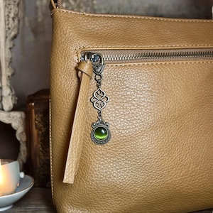 May include: A tan leather crossbody bag with a zipper closure and a decorative charm featuring a green gemstone. The charm is attached to the zipper pull.
