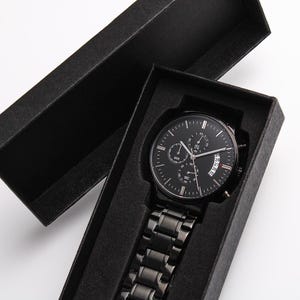 May include: A black wristwatch with a black metal band is displayed inside a black box. The watch face has multiple sub-dials and a date display. The watch is a sleek, modern accessory.