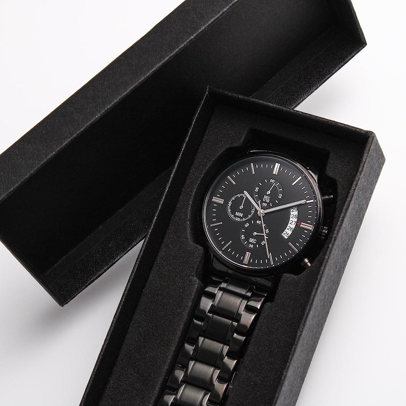 May include: A black wristwatch with a black metal band is displayed inside a black box. The watch face has multiple sub-dials and a date display. The watch is a sleek, modern accessory.