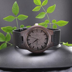 Pode incluir: Um rel&oacute;gio de pulso de madeira com mostrador castanho escuro e bracelete de couro preto. O rel&oacute;gio tem uma moldura prateada e uma pequena coroa prateada. O rel&oacute;gio &eacute; exibido numa superf&iacute;cie preta com folhas verdes no fundo.