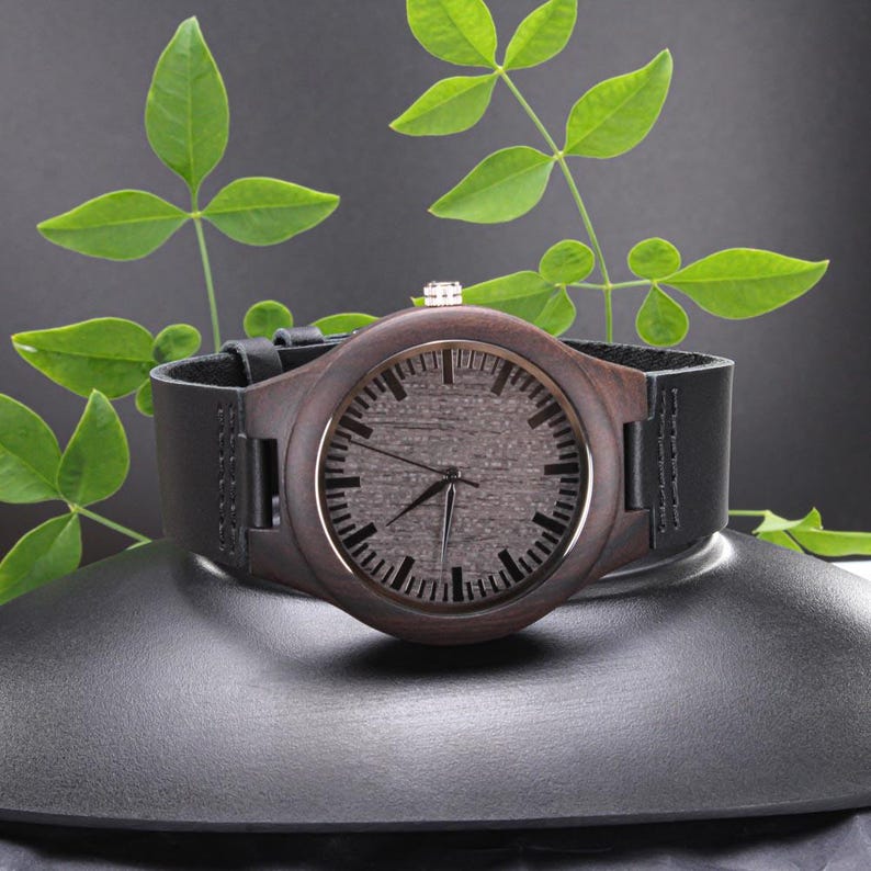 Pode incluir: Um rel&oacute;gio de pulso de madeira com mostrador castanho escuro e bracelete de couro preto. O rel&oacute;gio tem uma moldura prateada e uma pequena coroa prateada. O rel&oacute;gio &eacute; exibido numa superf&iacute;cie preta com folhas verdes no fundo.