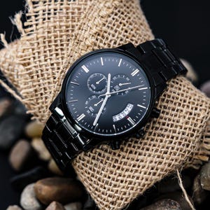 May include: A black wristwatch with a black metal band and a black face. The watch has multiple sub-dials and white hands. The watch is resting on a piece of burlap fabric and small rocks.