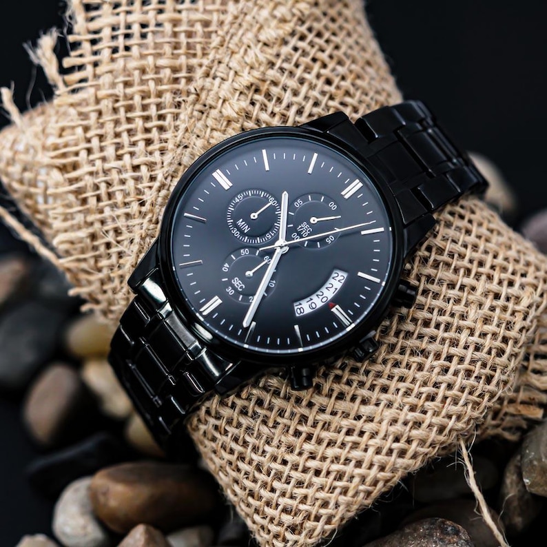 May include: A black wristwatch with a black metal band and a black face. The watch has multiple sub-dials and white hands. The watch is resting on a piece of burlap fabric and small rocks.