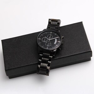 May include: A black chronograph watch with a matching black metal band rests on a black box. The watch face features multiple sub-dials and white hands. The watch is a sleek, modern accessory.