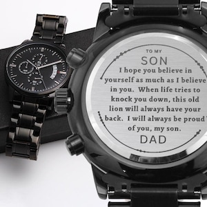 Può includere: Due orologi neri. Un orologio ha un quadrante nero con segni bianchi e un cinturino in metallo nero. L'altro orologio ha un quadrante nero con la scritta "TO MY SON" e un messaggio da "DAD".