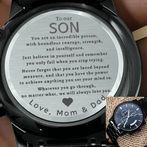 Puede incluir: Reloj de pulsera negro con esfera redonda. La esfera del reloj tiene un mensaje grabado: "To our SON" seguido de un mensaje inspirador de "Love, Mom & Dad". El reloj tiene una correa de metal negro y un bisel negro.