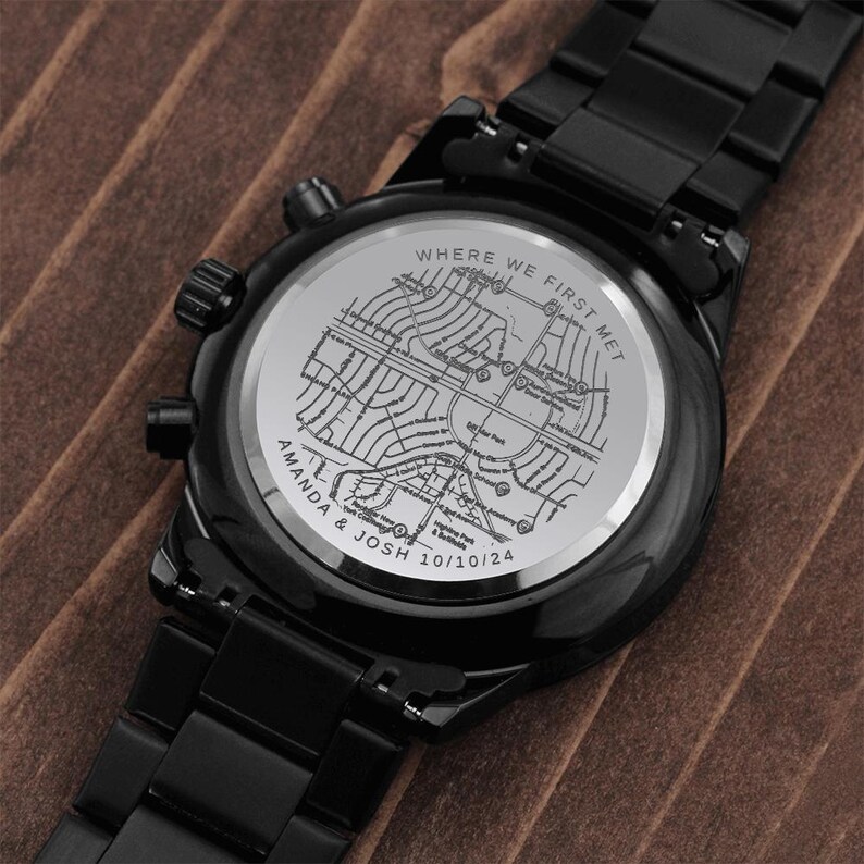 May include: Black wristwatch with a black metal band. The watch face features a map design with the text "WHERE WE FIRST MET" and the names "AMANDA & JOSH 10/10/24". The watch is displayed on a brown surface.