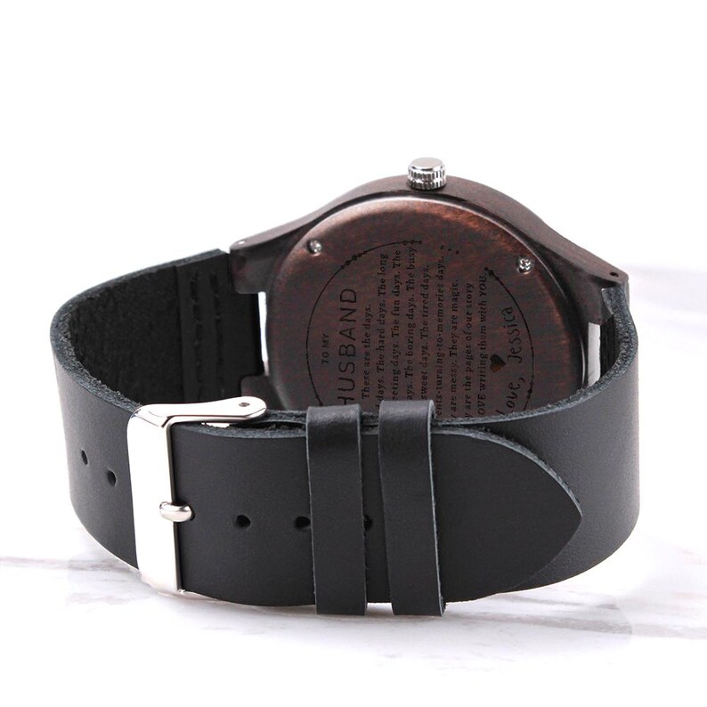 Pode incluir: Um rel&oacute;gio de pulso de madeira castanha escura com uma bracelete de couro preta e uma fivela prateada. O mostrador do rel&oacute;gio tem gravado o texto "TO MY HUSBAND" e uma mensagem.