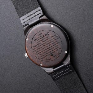 Pode incluir: Um rel&oacute;gio de pulso de madeira com uma pulseira de couro preta. O mostrador do rel&oacute;gio &eacute; gravado com uma mensagem para um marido, incluindo as palavras "Love, Jessica". O rel&oacute;gio tem um mostrador de madeira castanho escuro e uma coroa prateada.