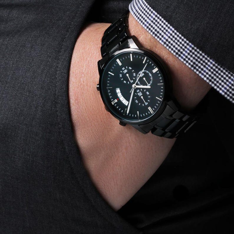 May include: A black wristwatch with a black metal band. The watch face is black with white markings and three sub-dials. The watch is worn on a wrist, with a dark suit jacket in the background.