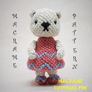 Puede incluir: Una muñeca de oso de ganchillo con un vestido rosa con detalles azules y zapatos a juego. El oso tiene cabeza y brazos blancos, con ojos y nariz negros. El texto en la imagen dice "MACRAME PATTERN" y "MACRAME TUTORIAL PDF".