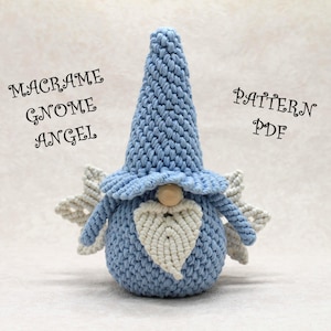 Puede incluir: Un ángel gnomo de macramé hecho a mano en azul claro con barba y alas blancas. El gnomo tiene un sombrero largo y puntiagudo y una nariz de madera. El texto "MACRAME GNOME ANGEL PATTERN PDF" se muestra en la imagen.