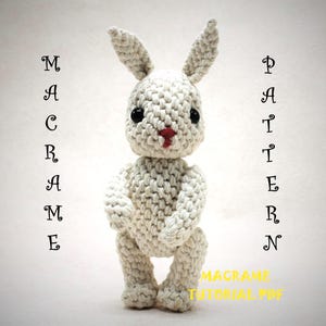 May include: A handcrafted, cream-coloured macrame rabbit with black eyes and a red nose. The rabbit is standing upright with its arms at its sides and its ears perked up. The words "MACRAME PATTERN" are written vertically on either side of the rabbit, with "MACRAME TUTORIAL PDF" at the bottom.