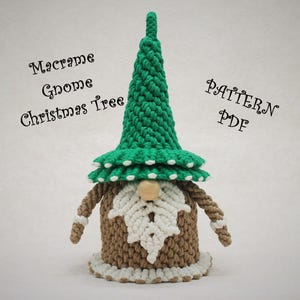 以下が含まれることがあります： 緑の尖った帽子と白いひげを持つ、手作りのマクラメノームクリスマスツリー。ノームは茶色の腕と体、木の鼻をしています。「Macrame Gnome Christmas Tree PATTERN PDF」の文字が見えます。