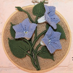 May include: A macrame wall hanging featuring three blue flowers with white centres and green leaves. The flowers are made with a knotting technique and are displayed in a wooden hoop.