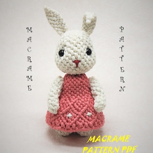 May include: A crocheted bunny toy, featuring a white body and a pink dress. The doll has black eyes and a pink nose. The image includes the text "MACRAME PATTERN PDF".