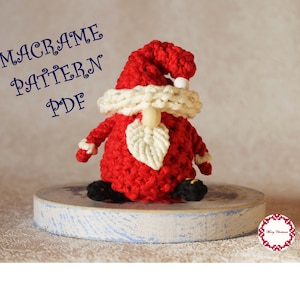 Könnte beinhalten: Ein roter und weißer gehäkelter Weihnachts-Gnome mit weißem Bart und einer Holznase. Der Gnome sitzt auf einem blauen und weißen Holzsockel. Der Text "MACRAME PATTERN PDF" befindet sich in der oberen linken Ecke des Bildes.