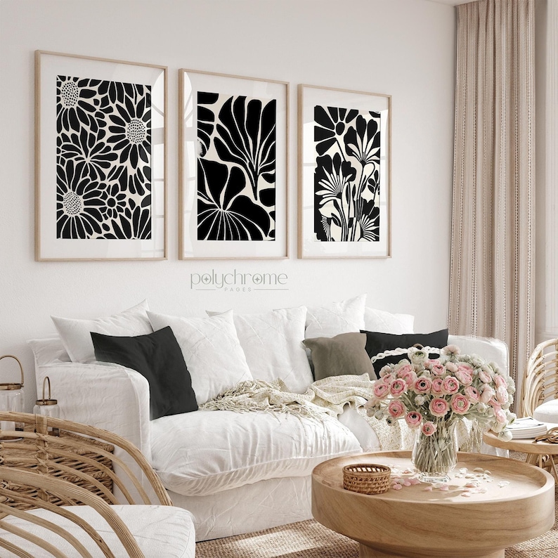 May include: Three black and white framed prints of stylized floral designs. The prints are hanging on a white wall above a white couch with black pillows. The couch is in a living room with a wooden coffee table and a wicker chair.