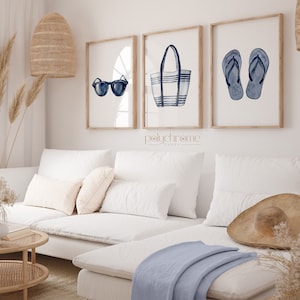 May include: Three framed watercolor illustrations of summer essentials: sunglasses, a striped tote bag, and flip flops. The illustrations are in shades of blue and white and are set against a white background. The frames are natural wood.