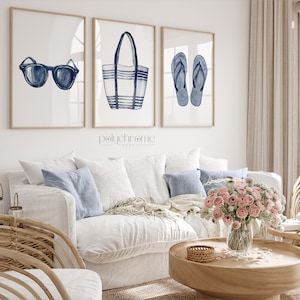 May include: Three watercolor illustrations of summer essentials: a pair of blue sunglasses, a striped blue and white beach bag, and a pair of blue flip flops. The illustrations are framed and displayed on a white wall above a white couch.