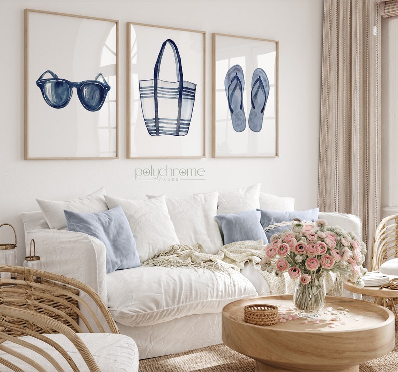 May include: Three watercolor illustrations of summer essentials: a pair of blue sunglasses, a striped blue and white beach bag, and a pair of blue flip flops. The illustrations are framed and displayed on a white wall above a white couch.