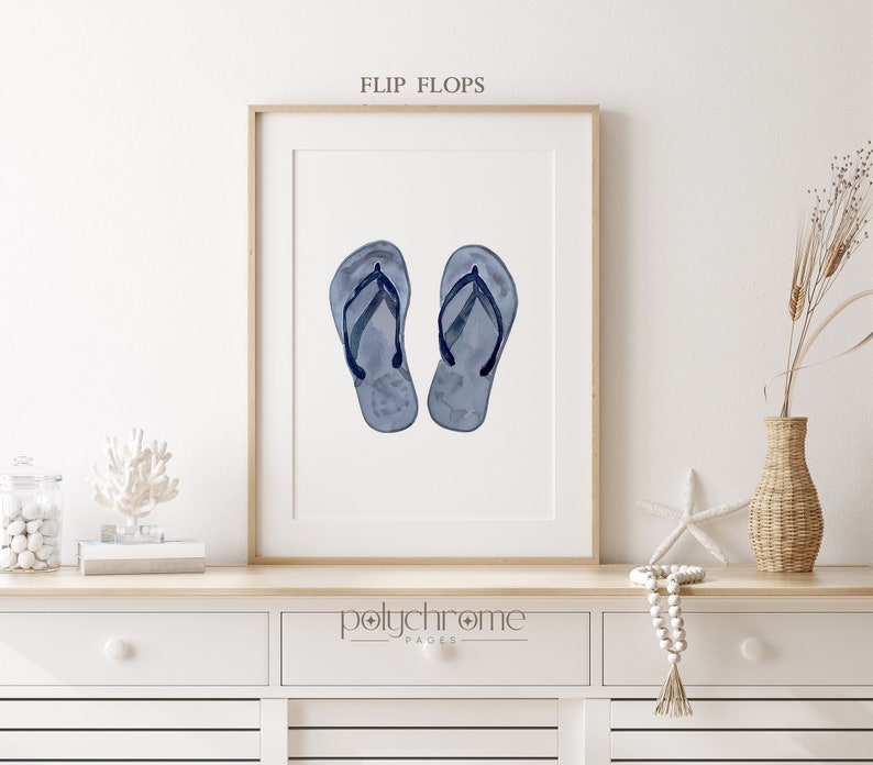 May include: A watercolor painting of a pair of blue flip flops with black straps. The painting is framed and displayed on a shelf with other beach-themed decor.