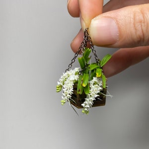 May include: A miniature hanging planter with white flowers and green leaves. The planter is made of wood and has a chain for hanging.
