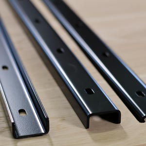 Op de afbeelding: Drie metalen plankdragers op een rij, elk met een U-vormig kanaal. De dragers zijn in tinten zilver en zwart, met diverse montagegaten. Ze liggen op een licht houten oppervlak.