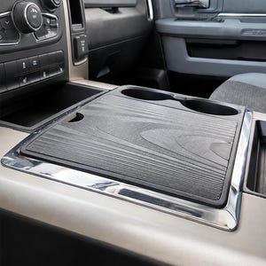 May include: A black centre console cover with a textured wood grain pattern and two integrated cup holders. The rectangular cover features a sleek, dark finish and is designed to fit over the existing console area of a vehicle interior.