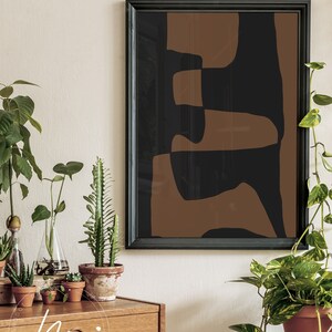 May include: A framed abstract art print with brown and black geometric shapes. The print is hanging on a white wall above a wooden dresser with plants.