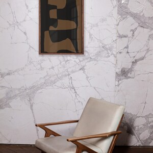 May include: A framed abstract art print with brown and black geometric shapes against a white marble wall. A white armchair with wooden arms sits in front of the wall.