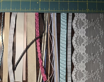 May include: Assortment of ribbons and lace trim in various colors and patterns, including beige, pink, blue, black, and white. The trim is arranged vertically on a green cutting mat.