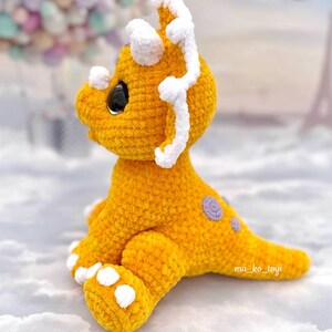 May include: A crocheted yellow triceratops dinosaur toy with white accents on the horns and feet. The dinosaur has purple spots on its back and a black eye.