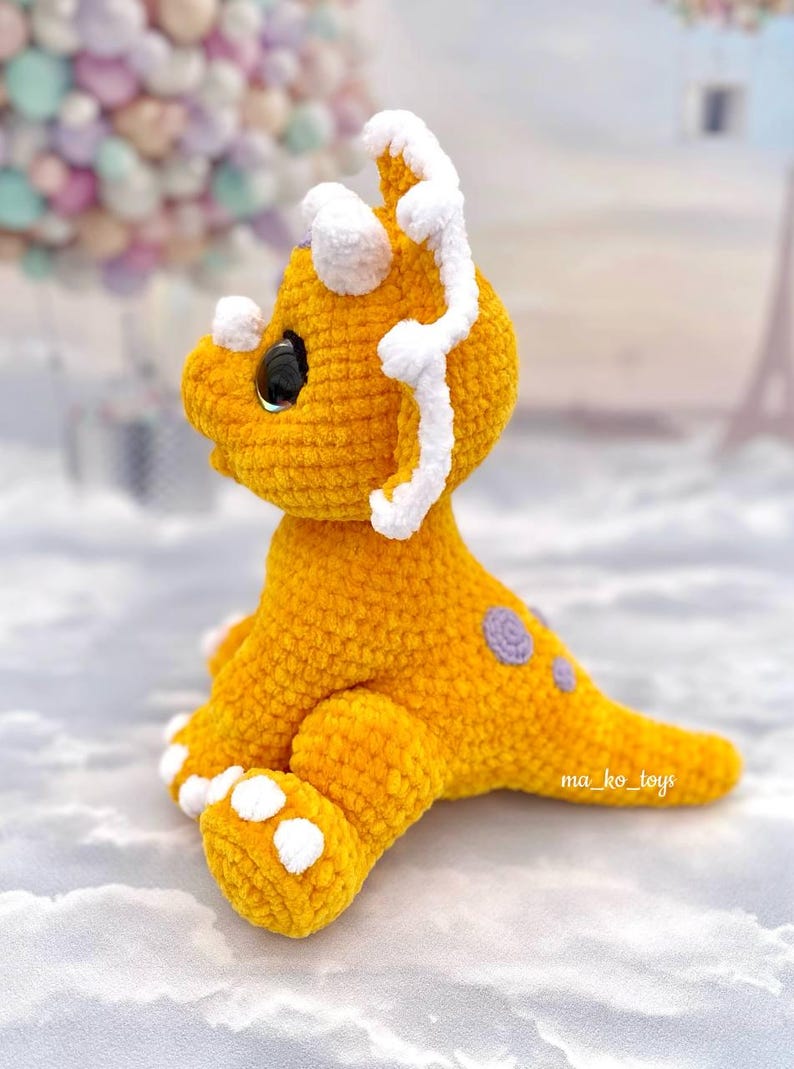 May include: A crocheted yellow triceratops dinosaur toy with white accents on the horns and feet. The dinosaur has purple spots on its back and a black eye.