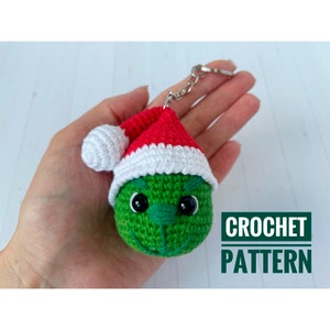 May include: A green crocheted character keychain wearing a red and white Santa hat. The keychain has black eyes and a stitched mouth. The text "CROCHET PATTERN" is in a green and white box.