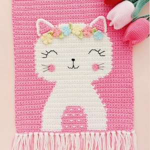 Peut inclure: Une tapisserie murale en crochet rose avec un chat blanc portant une couronne de fleurs. Le chat a un visage souriant et des joues roses. La tapisserie murale a une bordure en franges.