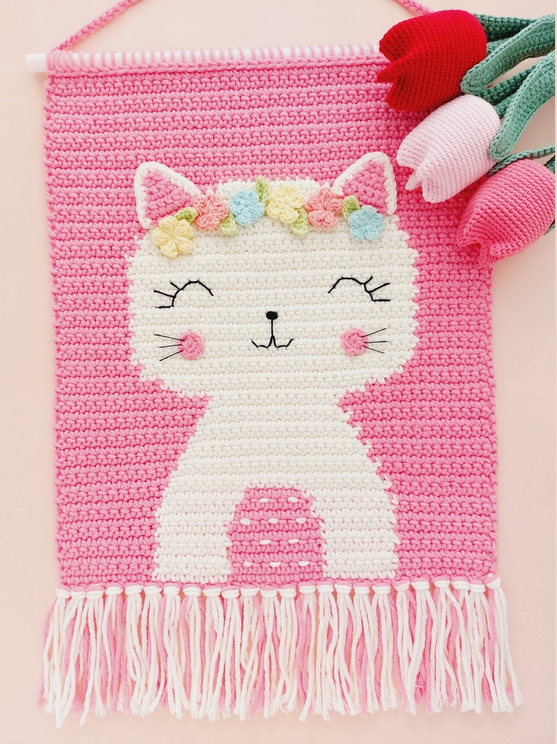 Peut inclure: Une tapisserie murale en crochet rose avec un chat blanc portant une couronne de fleurs. Le chat a un visage souriant et des joues roses. La tapisserie murale a une bordure en franges.