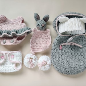 Pu&ograve; includere: Un set di vestiti per neonati all'uncinetto in rosa e grigio, tra cui un vestito, un copri pannolino, scarpine, un sacco a pelo con cappuccio e un sonaglio a forma di coniglio.