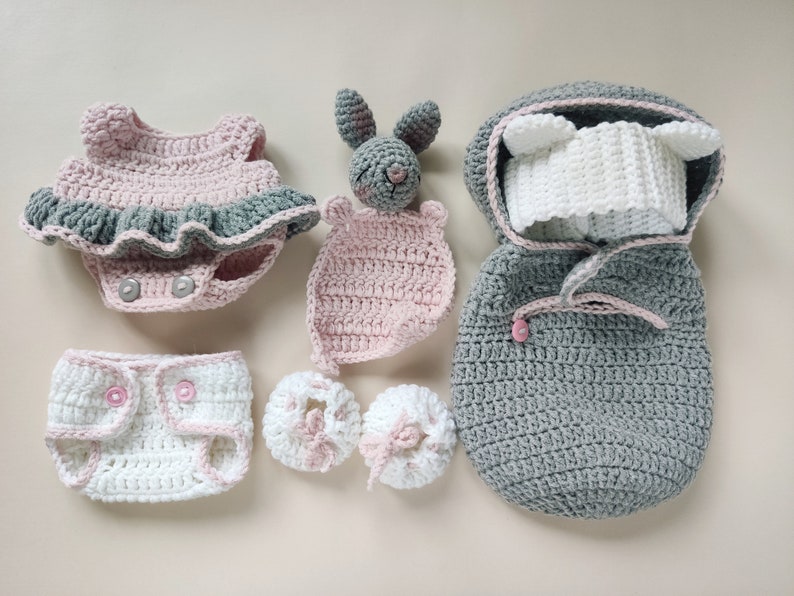 Pu&ograve; includere: Un set di vestiti per neonati all'uncinetto in rosa e grigio, tra cui un vestito, un copri pannolino, scarpine, un sacco a pelo con cappuccio e un sonaglio a forma di coniglio.