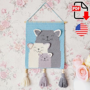May include: A handmade crochet wall hanging featuring a family of cats. The design includes a blue background, with three cats in shades of gray, white, and lavender. The piece is adorned with tassels and wooden beads, and is suspended from a wooden dowel.