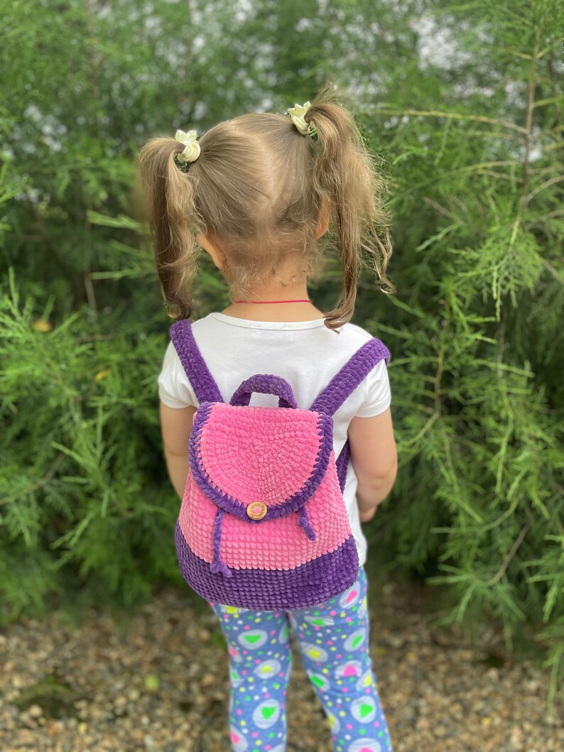 Pu&ograve; includere: Un bambino piccolo che indossa una camicia bianca e leggings colorati indossa uno zaino in crochet rosa e viola. Lo zaino ha un piccolo bottone di legno.