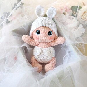 Pu&ograve; includere: Una bambola di bambino in crochet con un cappello bianco e orecchie di coniglio, che indossa un pannolino in crochet bianco, seduta in una scatola di tessuto di tulle bianco.