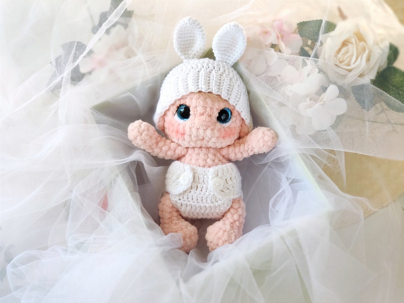 Pu&ograve; includere: Una bambola di bambino in crochet con un cappello bianco e orecchie di coniglio, che indossa un pannolino in crochet bianco, seduta in una scatola di tessuto di tulle bianco.