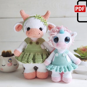 Può includere: Due animali di peluche all'uncinetto, una mucca e un unicorno, entrambi con abiti verdi e blu con bottoni bianchi. La mucca ha il naso rosa e una corona di fiori verdi. L'unicorno ha un corno rosa e occhi blu.