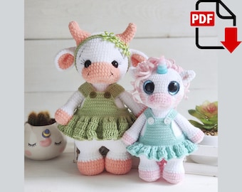 Puede incluir: Dos animales de peluche tejidos a crochet, una vaca y un unicornio, ambos con vestidos verdes y azules con botones blancos. La vaca tiene la nariz rosa y una corona de flores verdes. El unicornio tiene un cuerno rosa y ojos azules.